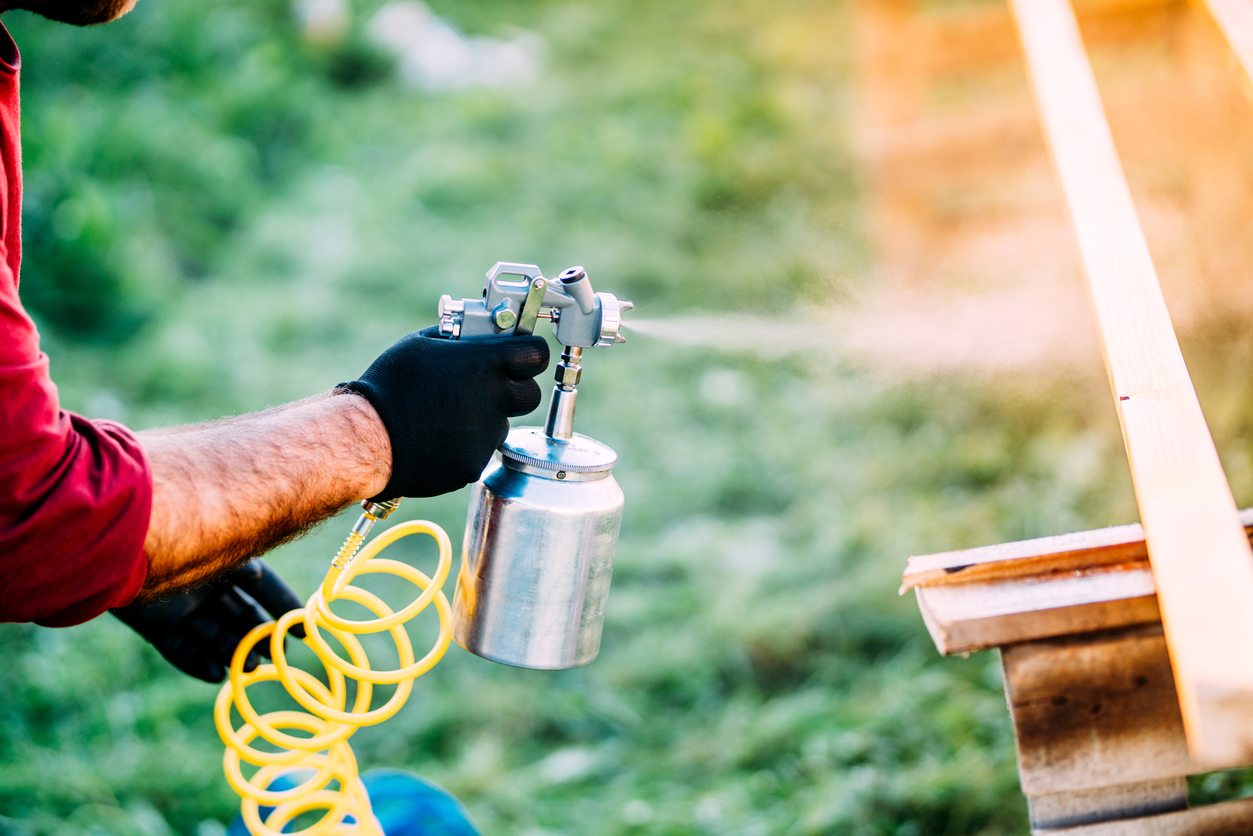 A man uses a tool connected to an air compressor to paint or sand-blast a board on a sawhorse.