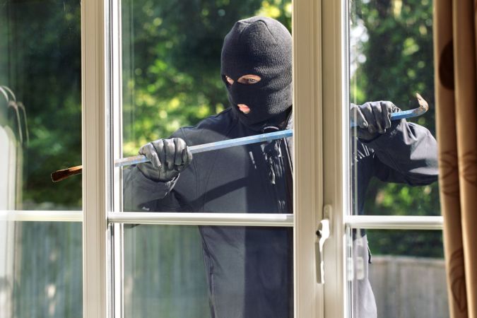 A burglar in a black ski mask is seen breaking into a home through a window.