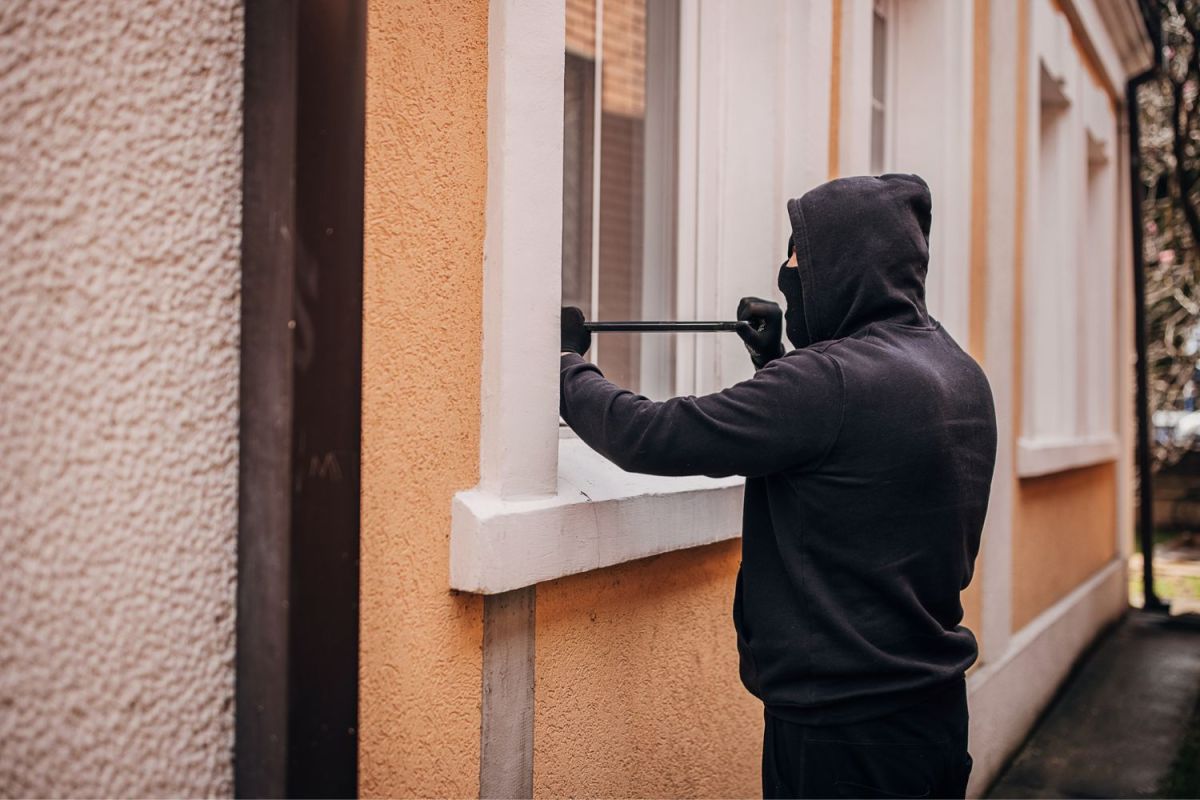 A burglar dressed in black uses a tool to break into a building's window.