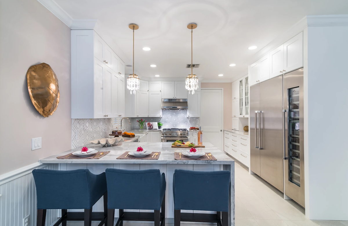 A predominantly white kitchen features stainless steel appliances, three navy blue chairs at the kitchen counter, and a golden metal wall art piece.