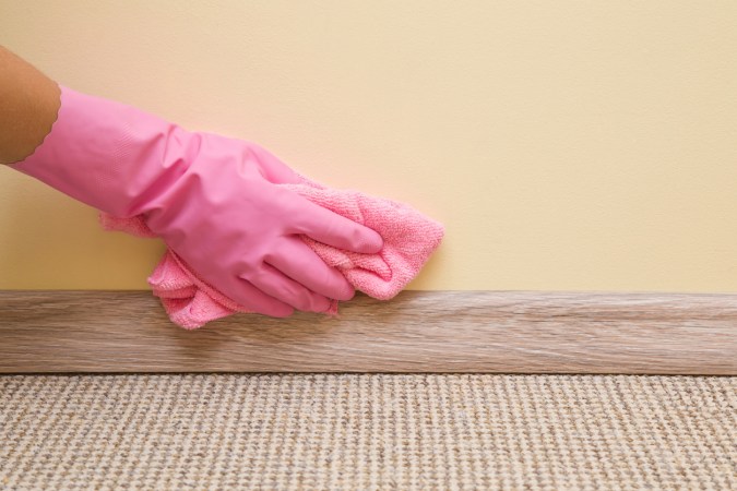 A person wearing a pink rubber glove wiping above the baseboard of a yellow wall with a flat paint texture.