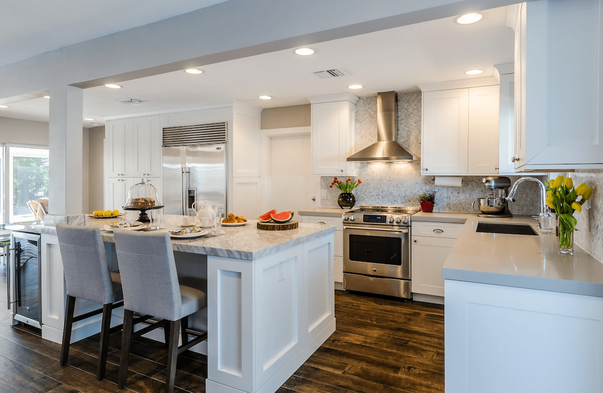 A white kitchen features dark hardwood flooring, plenty of marble counter space, and a refrigerator within the kitchen island.