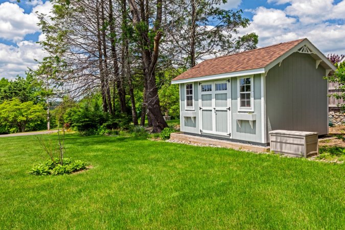 A grey shed sits on a lawn with tress in the background.