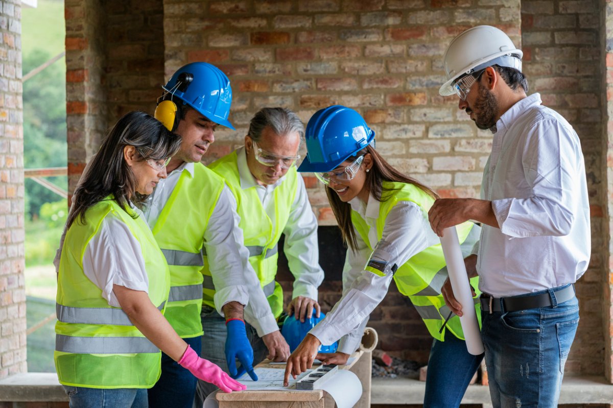 Five workers in hard hats and yellow safety vests consult a building plan. 