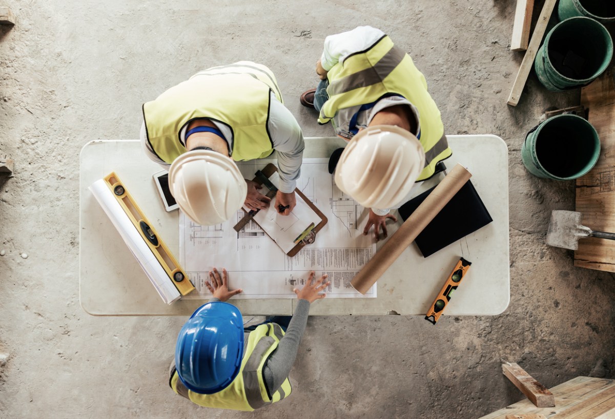 An aerial shot of three workers in hard hats consulting a building plan in an area under construction.