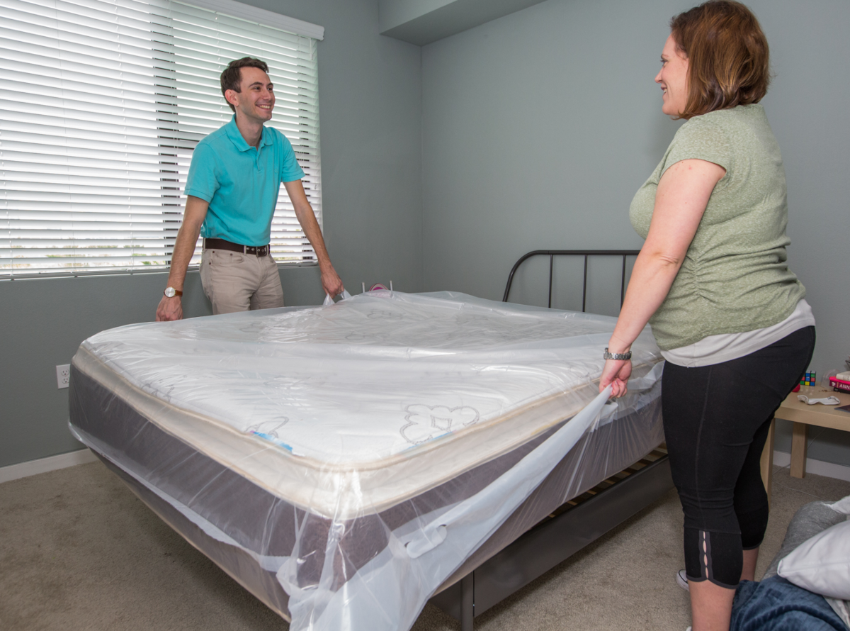 A man and woman are standing on either side of a covered mattress in a bedroom while preparing to lift it by the handles of the mattress cover.