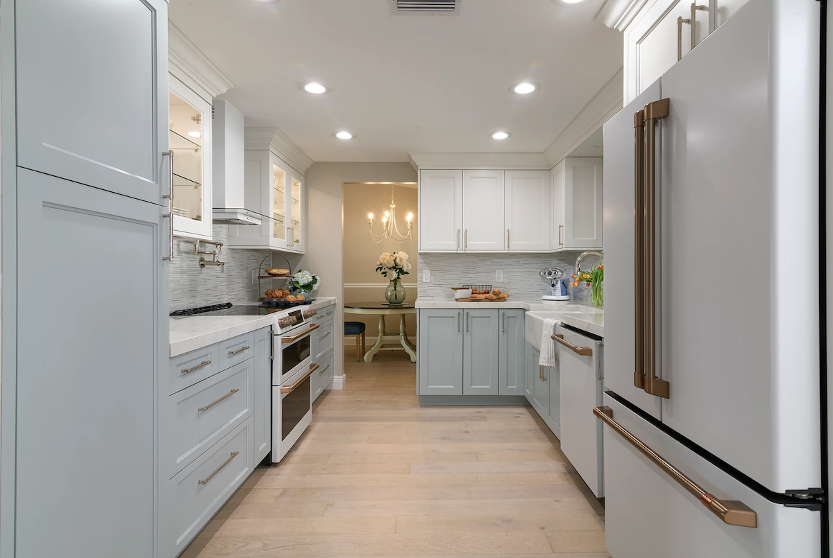 A coastal-themed kitchen features light wood flooring, white and light blue cabinetry, and white appliances with copper handles.