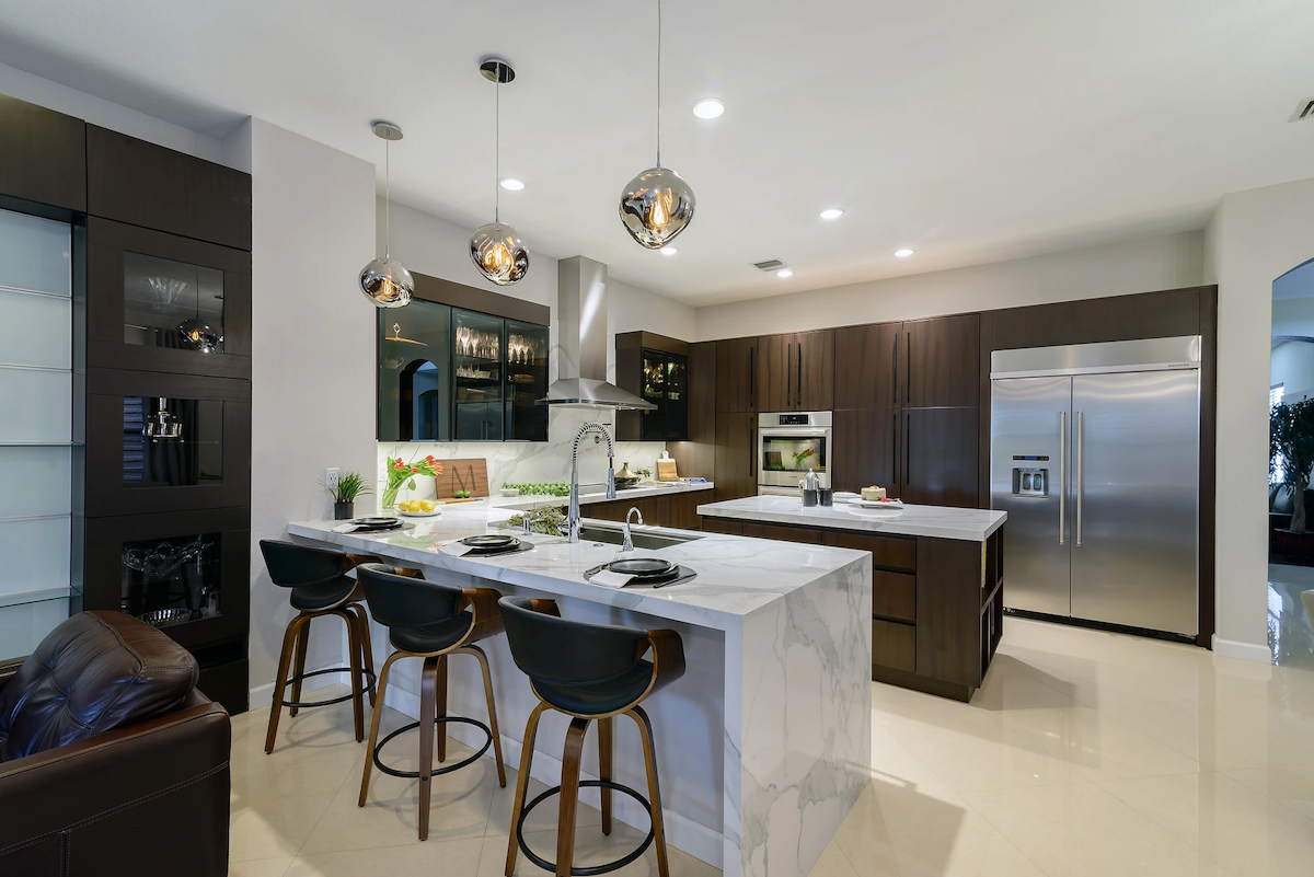 A remodeled kitchen features brown slab cabinets, white marble counters, bar stools, and trendy pendant lighting.