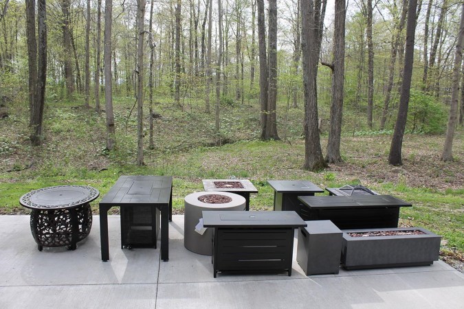 A group of the best fire pit tables on a cement patio near a wooded area.