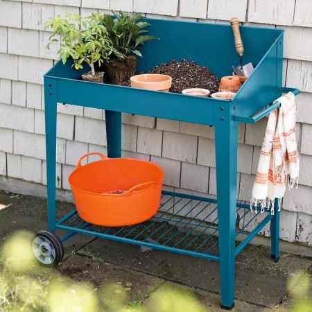  The Demeter Rolling Metal Potting Bench with supplies and plants next to the side of a house.