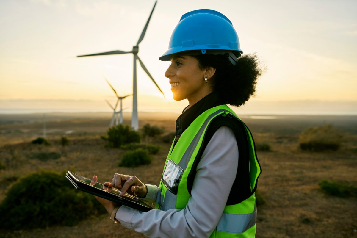 A worker in a blue hard hat smiles as wind turbines spin behind her. 