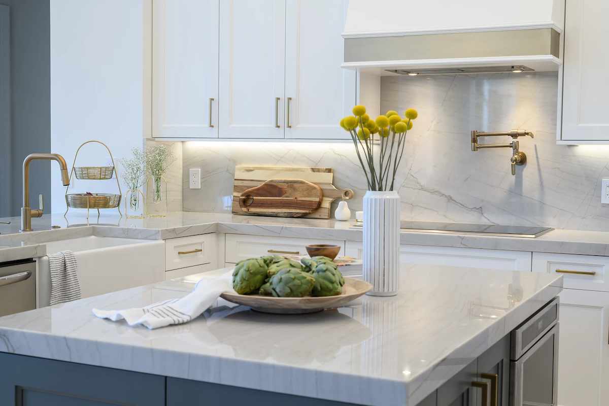 A white kitchen features a small kitchen island with quartzite countertops, lighting underneath the cabinets, and warm gold hardware.