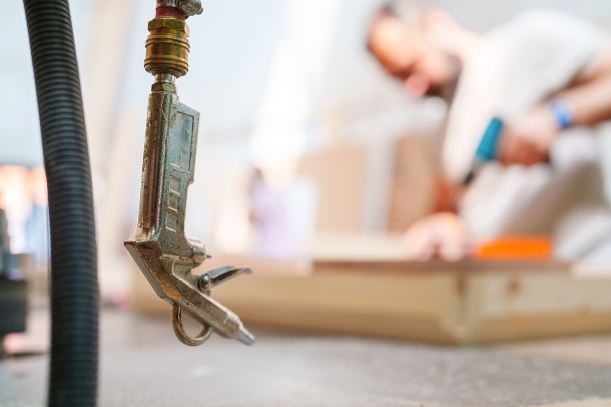 An in-focus air compressor hose hangs in front of an out-of-focus man using a power drill in a home workshop.