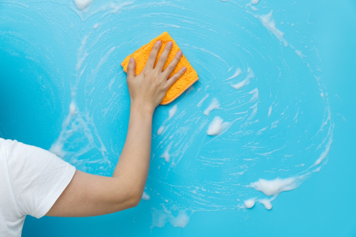 A person washes a flat-textured blue wall with a soapy microfiber cloth.