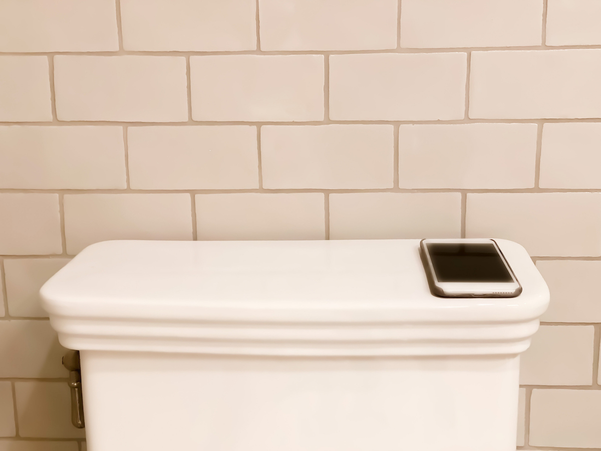 Smart phone resting on the back of a white toilet tank with white subway tile in the background.