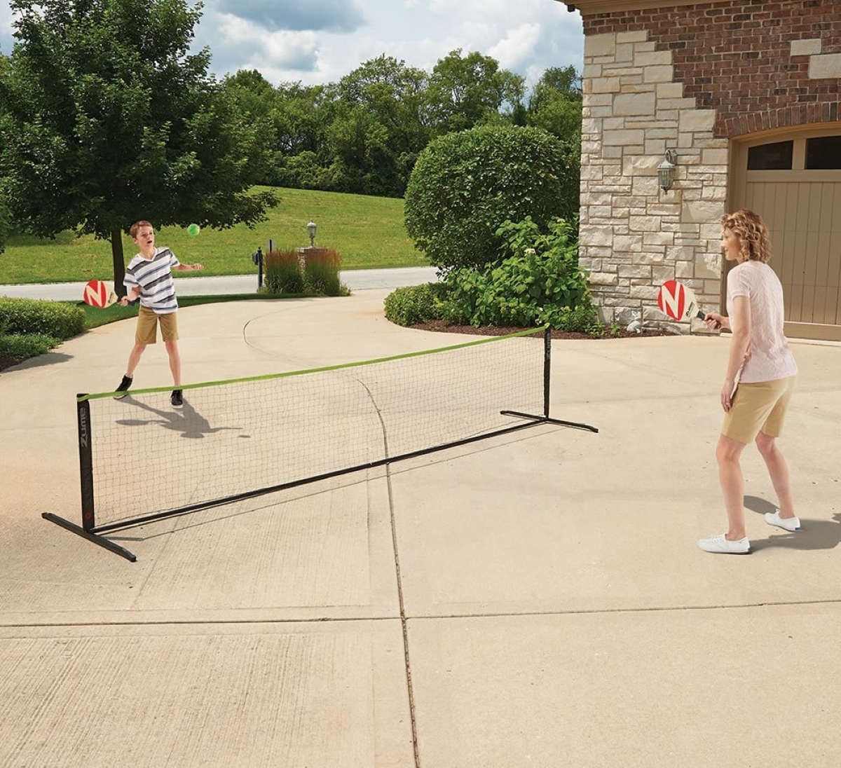 Two people are playing pickleball over a net in their driveway.