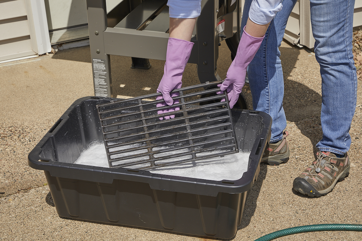 Woman wearing rubber gloves soaks a grill grate in a basin of soapy water, outdoors in front of a gas grill.