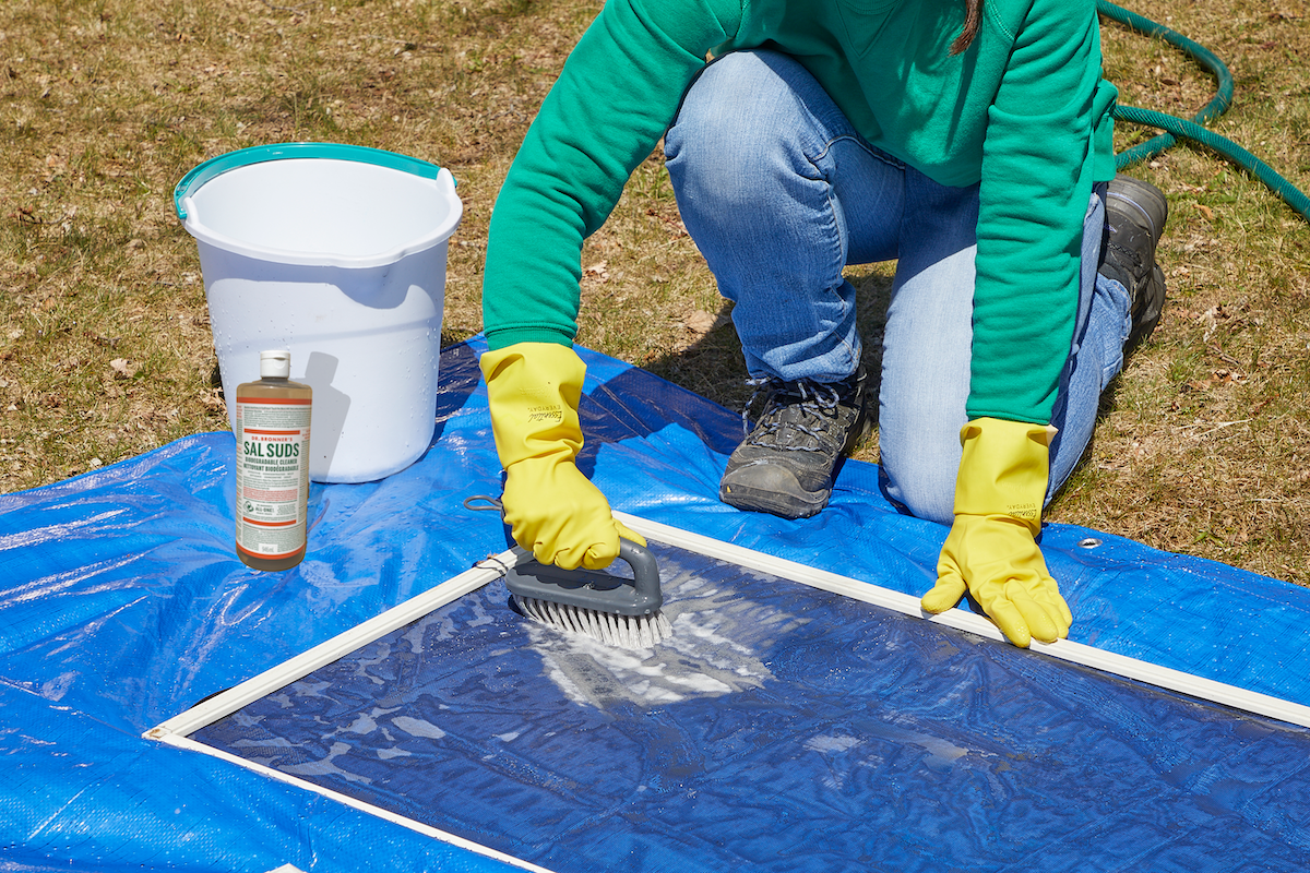 Woman scrubs a window screen on a blue tarp with a scrub brush.