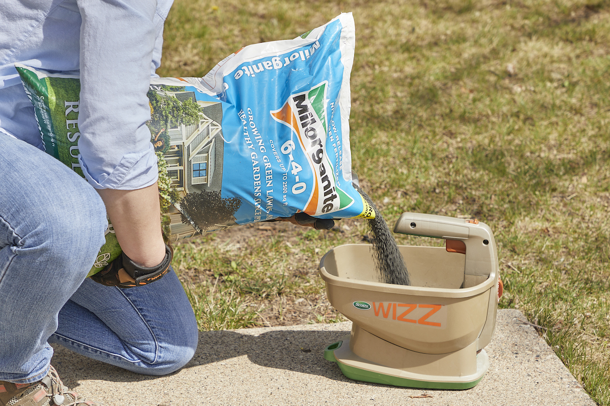 Woman pours fertilizer from a bag into a seed spreader for distribution on her grass.