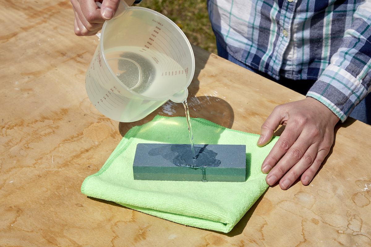 Woman using a measuring cup to pour water onto a sharpening stone atop a green microfiber cloth.