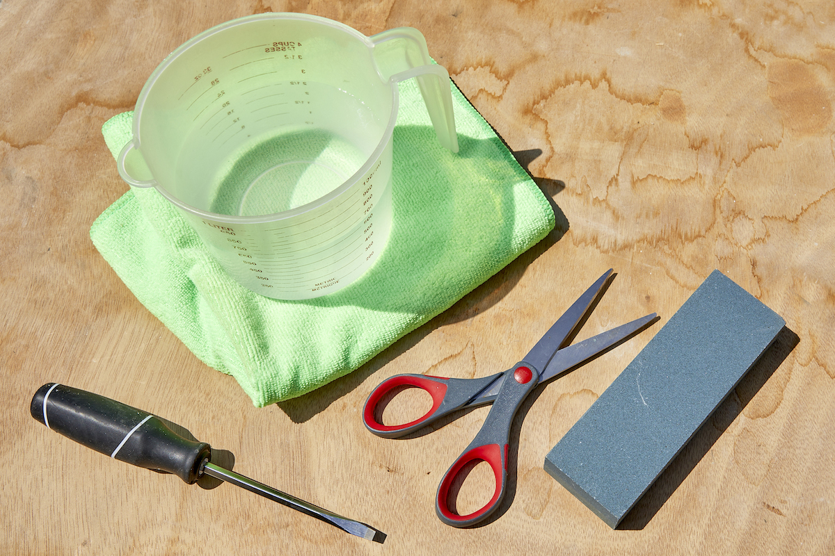 Materials needed to sharpen scissors, including sharpening stone, scissors, screwdriver, and cup of water.