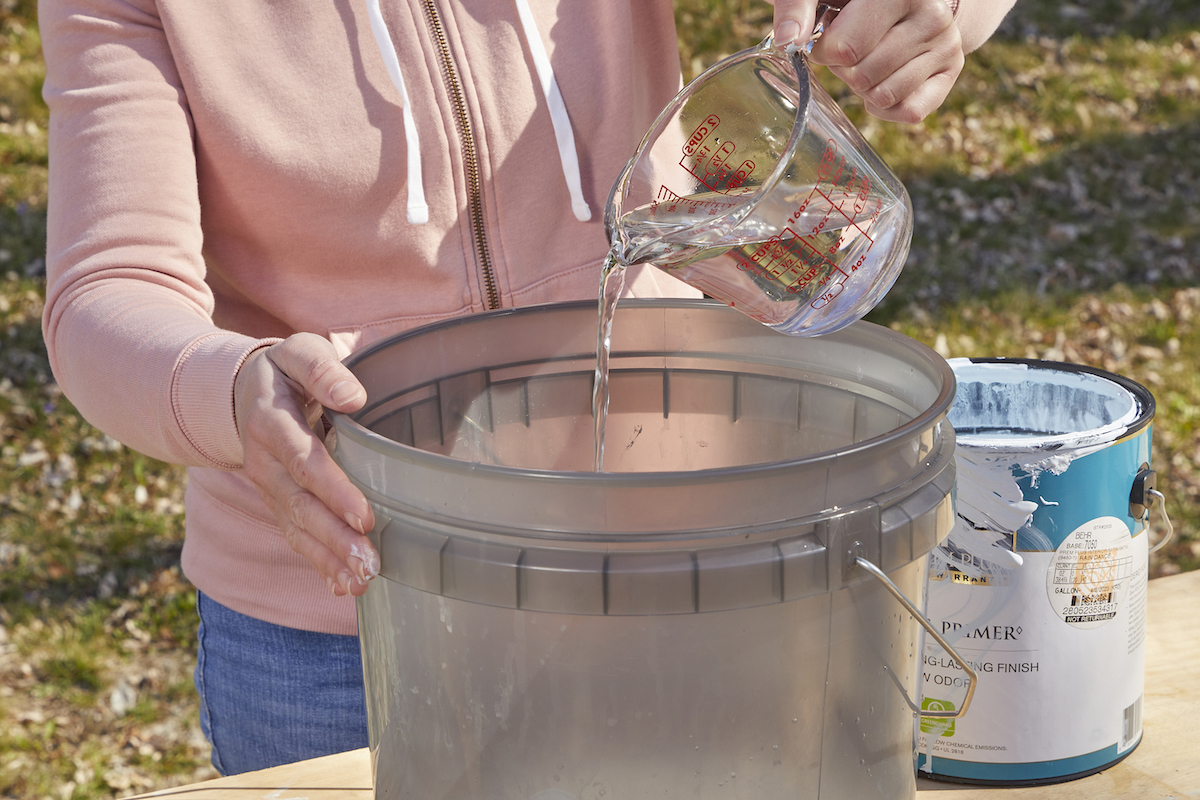 Woman pours water from a Pyrex measuring cup into a large bucket with white paint.