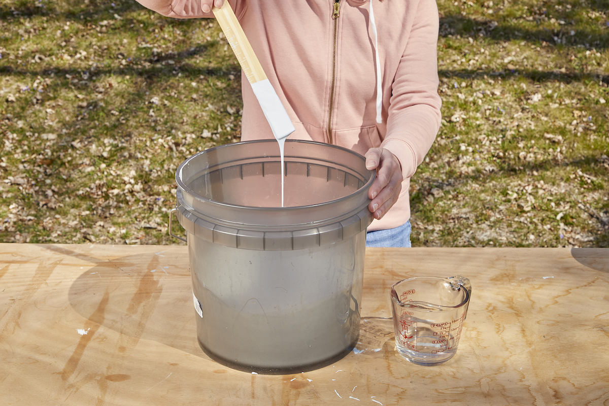 Woman holds a paint stir stick dripping with white paint above a bucket to evaluate the paint's thickness.