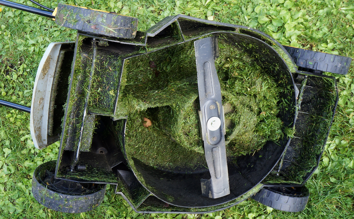 An electric lawn mower is upside down on a lawn, displaying wet grass clogging the blades.