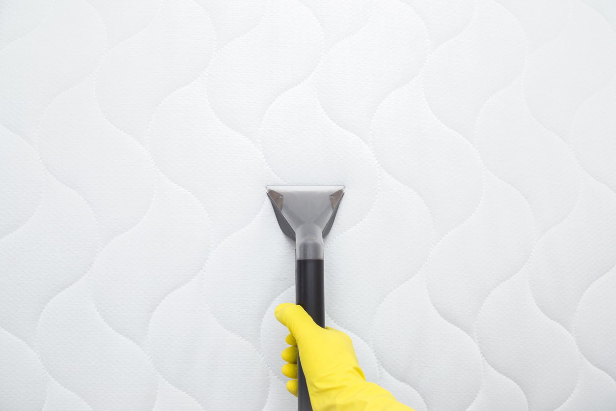 A person wearing yellow cleaning gloves uses a vacuum attachment to vacuum a white mattress.