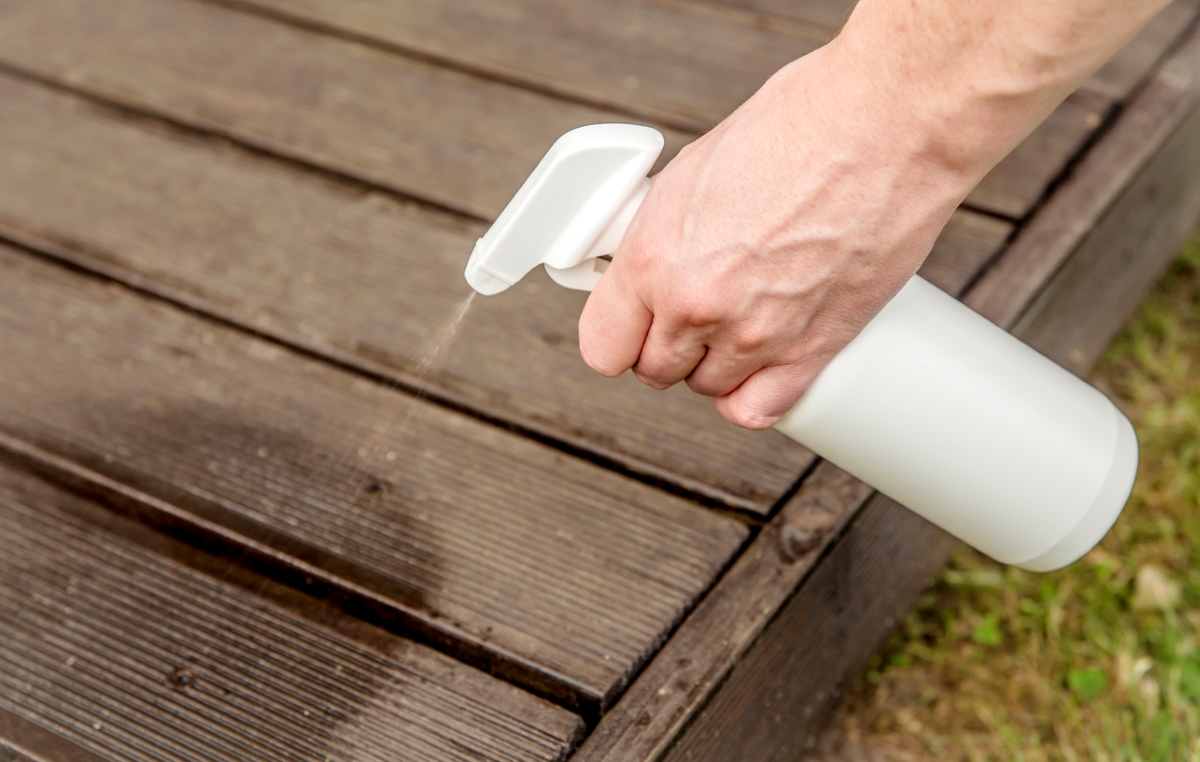 A person spraying a wooden deck with a clear substance using a white spray bottle.
