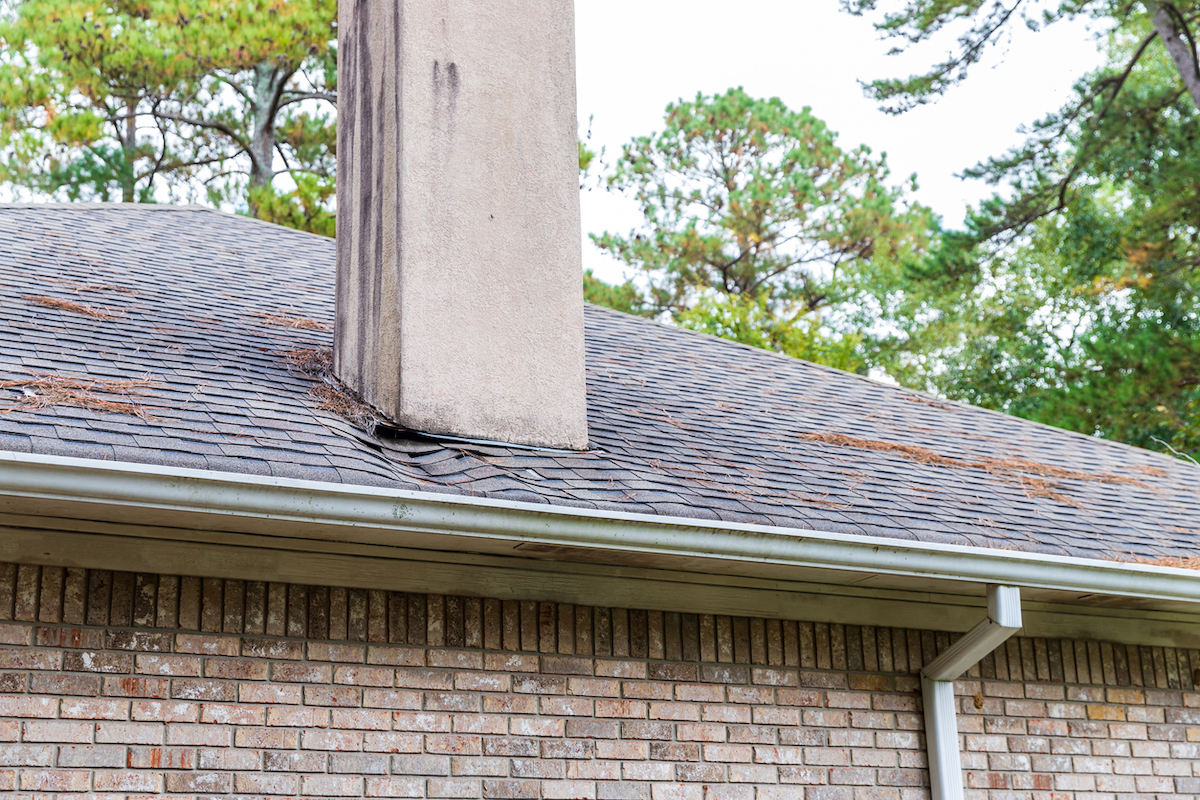 A house roof's shingles next to the chimney are warped from water damage.