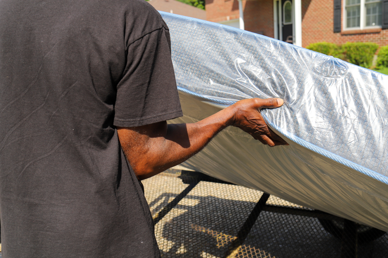 A person is lifting a covered mattress off of a trailer outdoors.