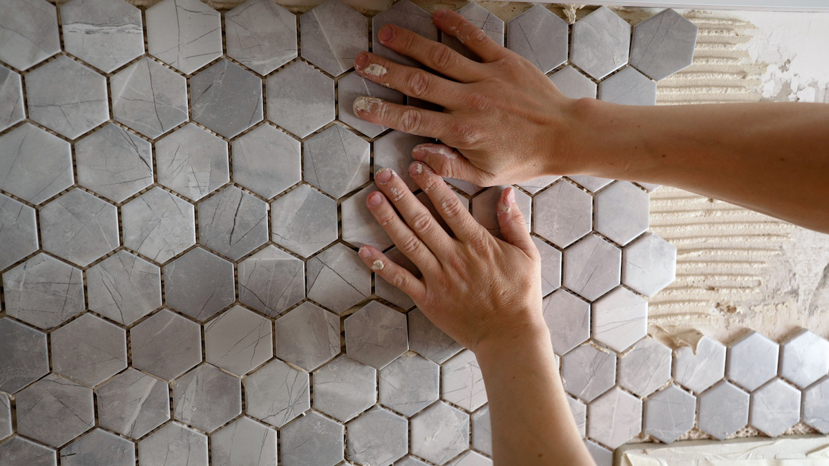 A person is laying grey hexagonal tile onto a bathroom wall.