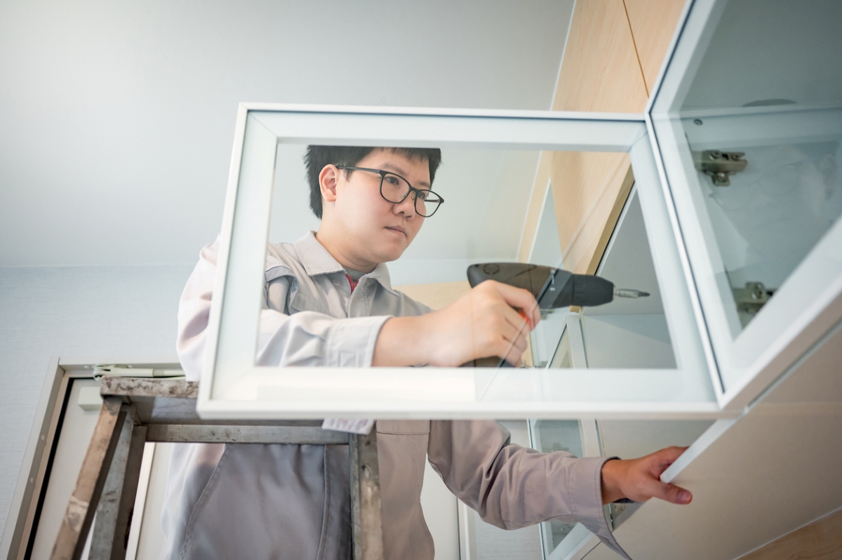 An Asian man on a ladder is using a drill to install a new clear kitchen cabinet door.
