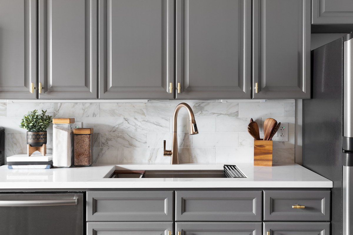A kitchen features grey cabinetry, white countertops, white marble backsplash, and gold hardware.