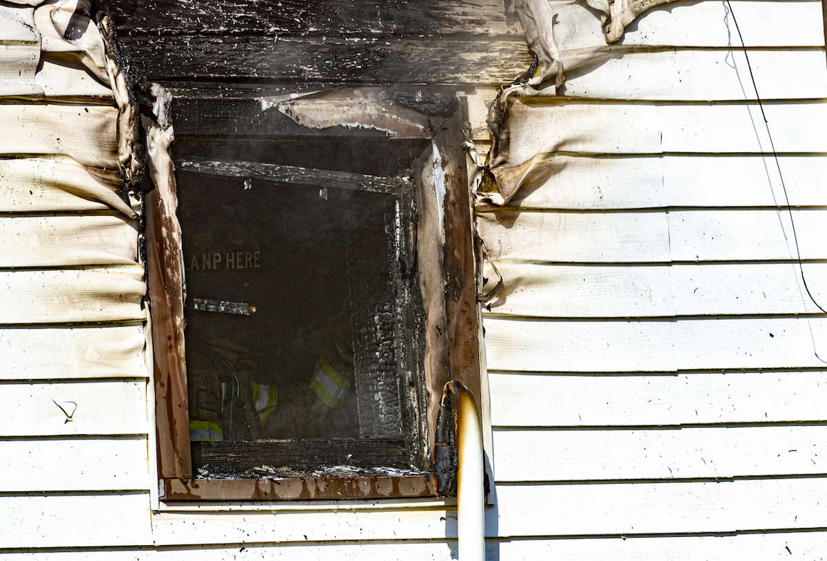 The siding and window of a white house is damaged by a fire that has been put out by the firefighters in the window.