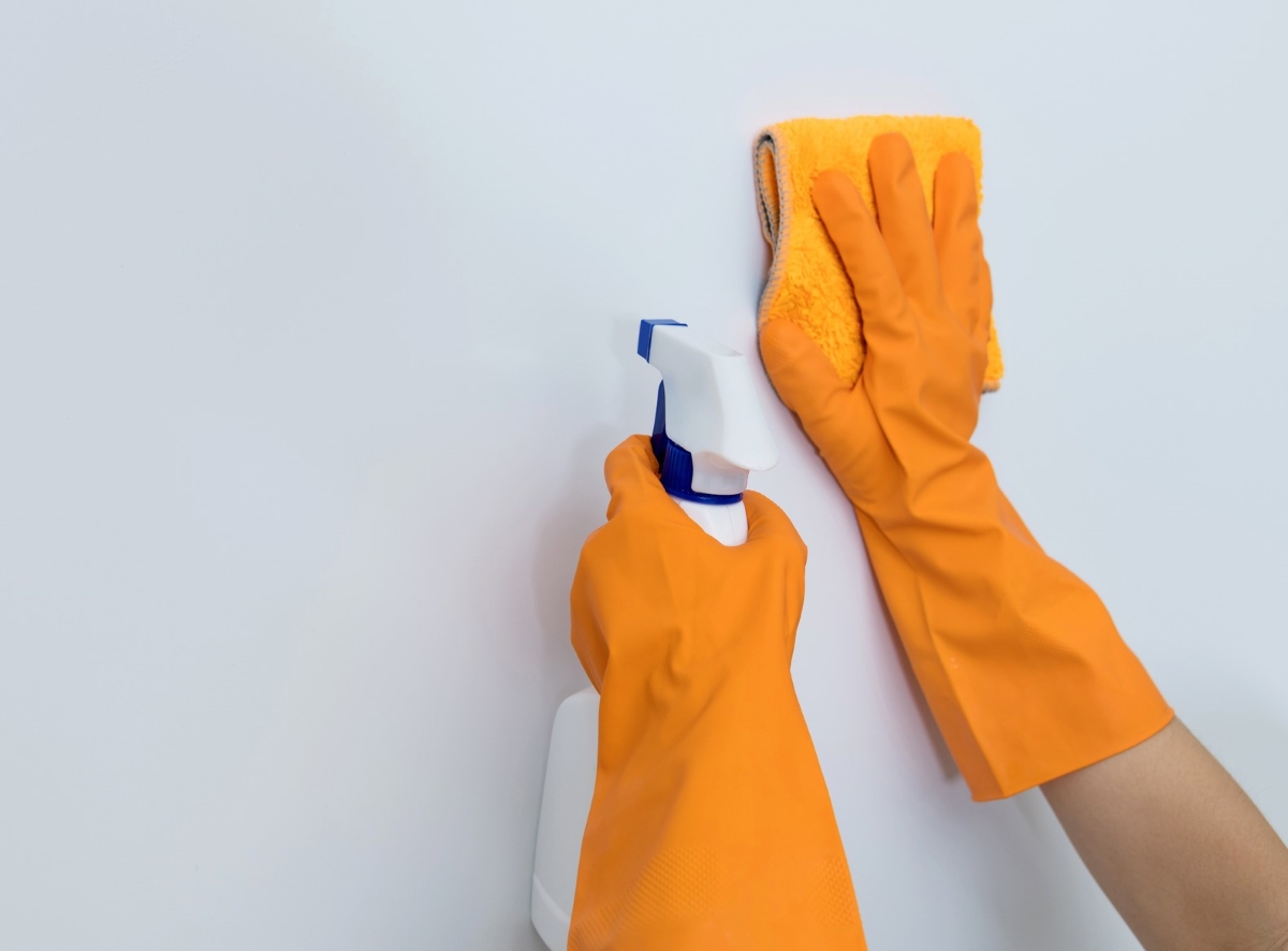 Orange gloved hands are cleaning a white wall with a rag and spray cleaner.