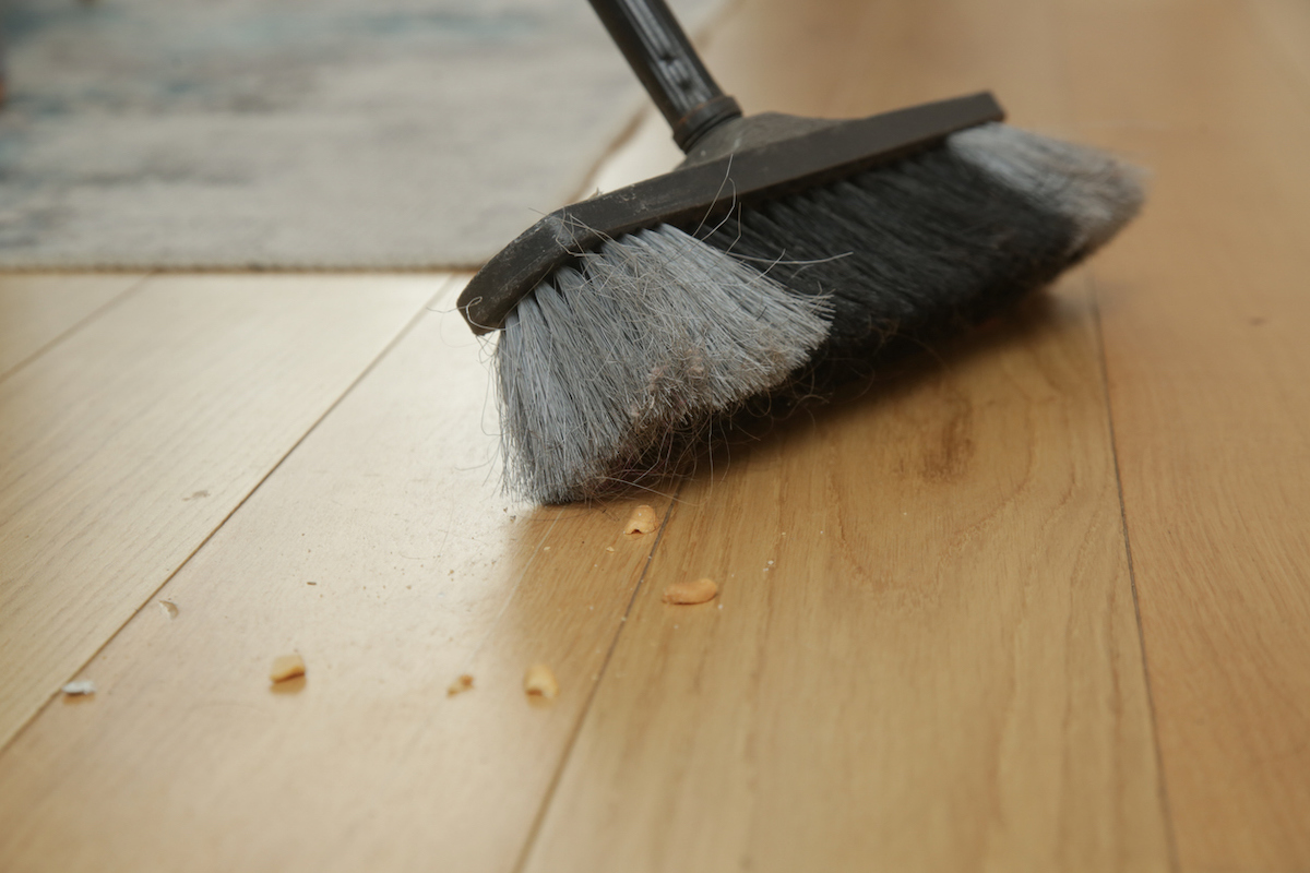 A black and gray broom is being used to sweep up food crumbs on hardwood flooring.