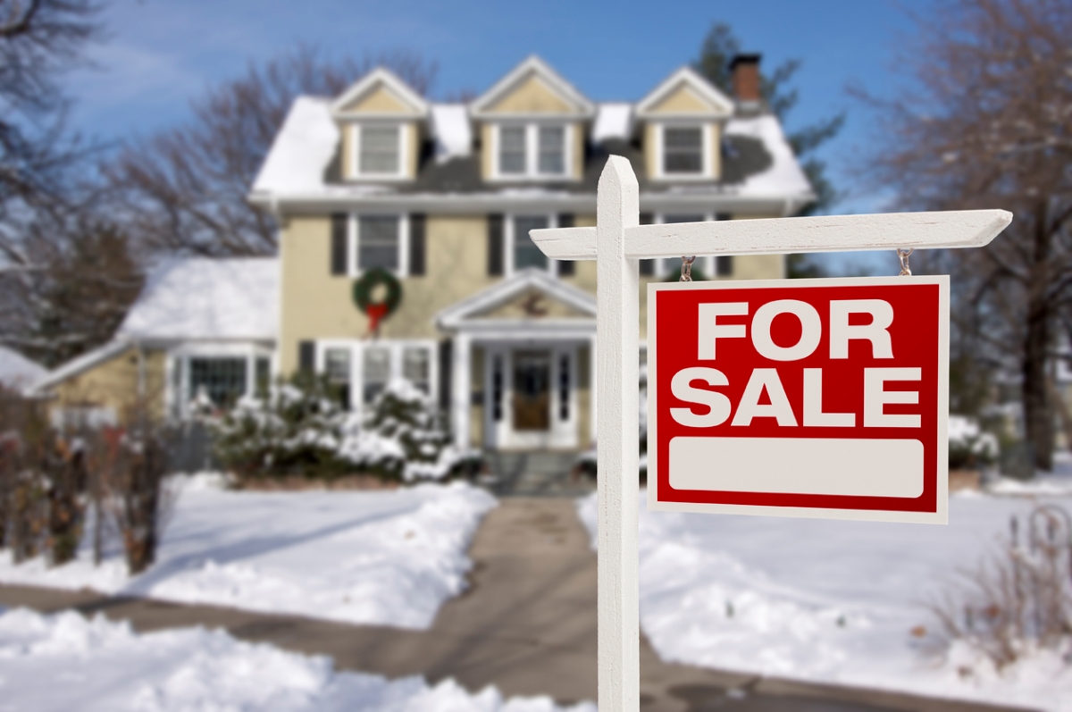 A for sale sign is placed in front of a home covered in snow.