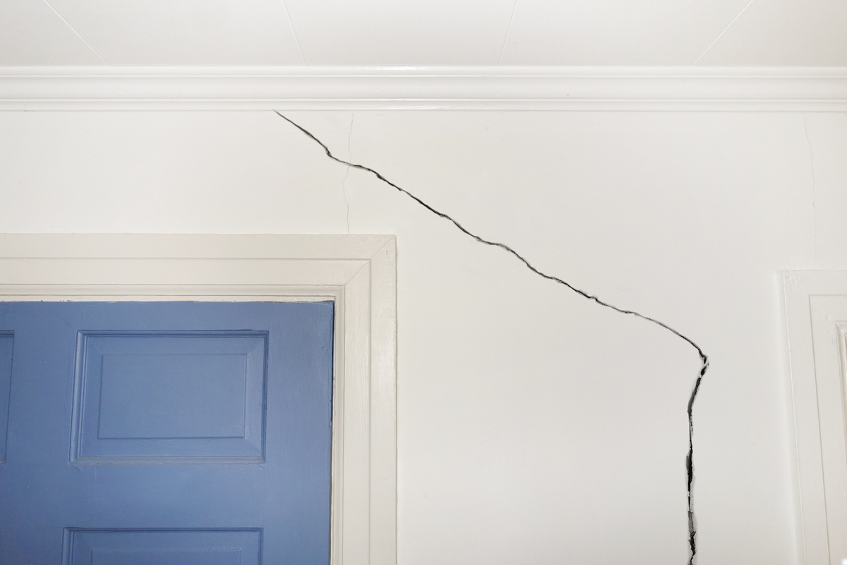 A crack in the wall of a home starts at the ceiling and continues next to the blue door.