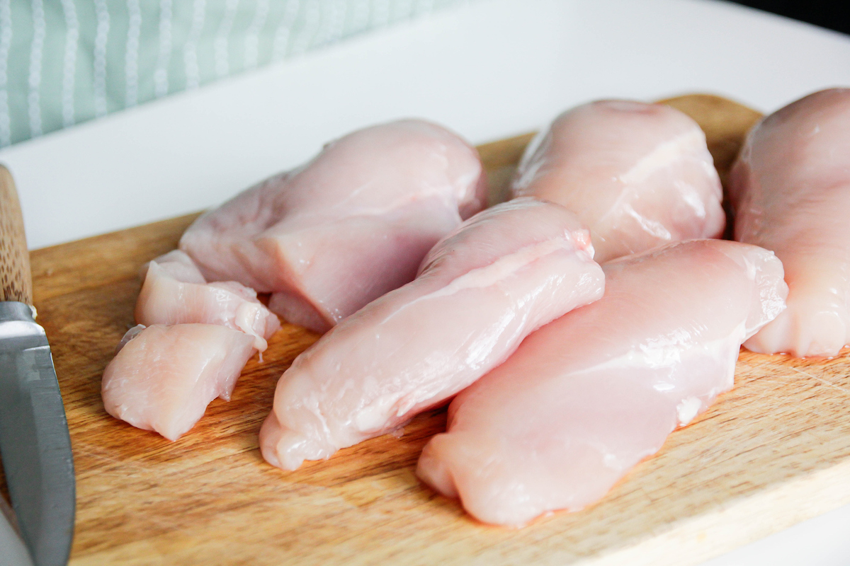 Five raw chicken breasts on a wooden cutting board with a knife nearby.