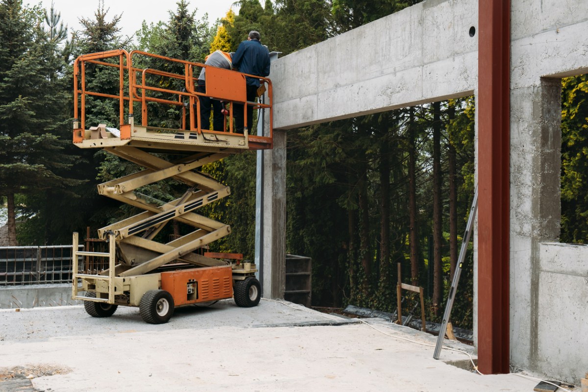 An orange scissor lift rental is raised next to a concrete wall.