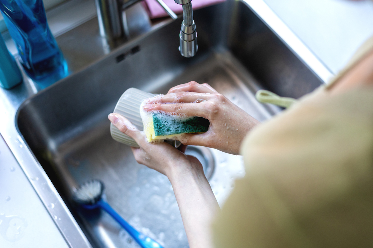 Woman washing a white mug at a kitchen sink with a scrub sponge.