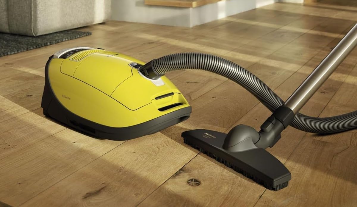A Miele Complete C3 Calima Canister Vacuum is resting on a hardwood flooring with sunlight shining on it.