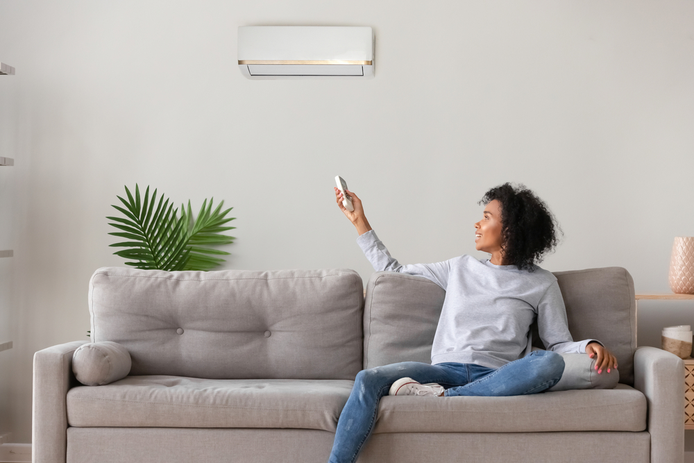 A woman sits on a couch and uses a remote to control a mounted AC over her head. 