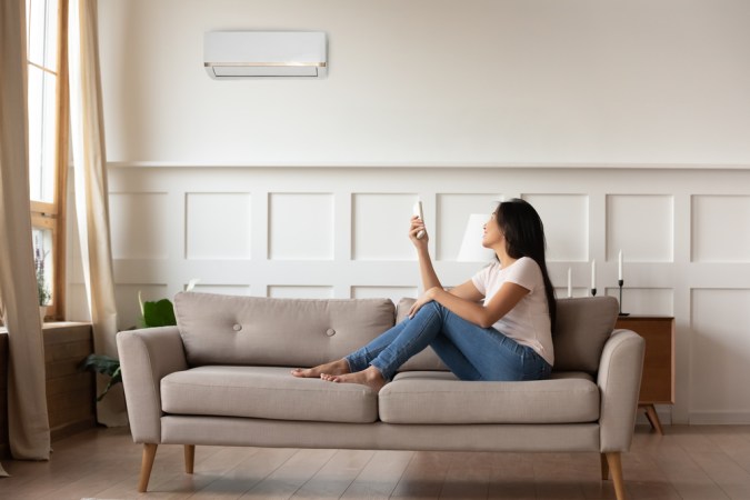 A young woman sits on a sofa, using a remote to manage an AC.