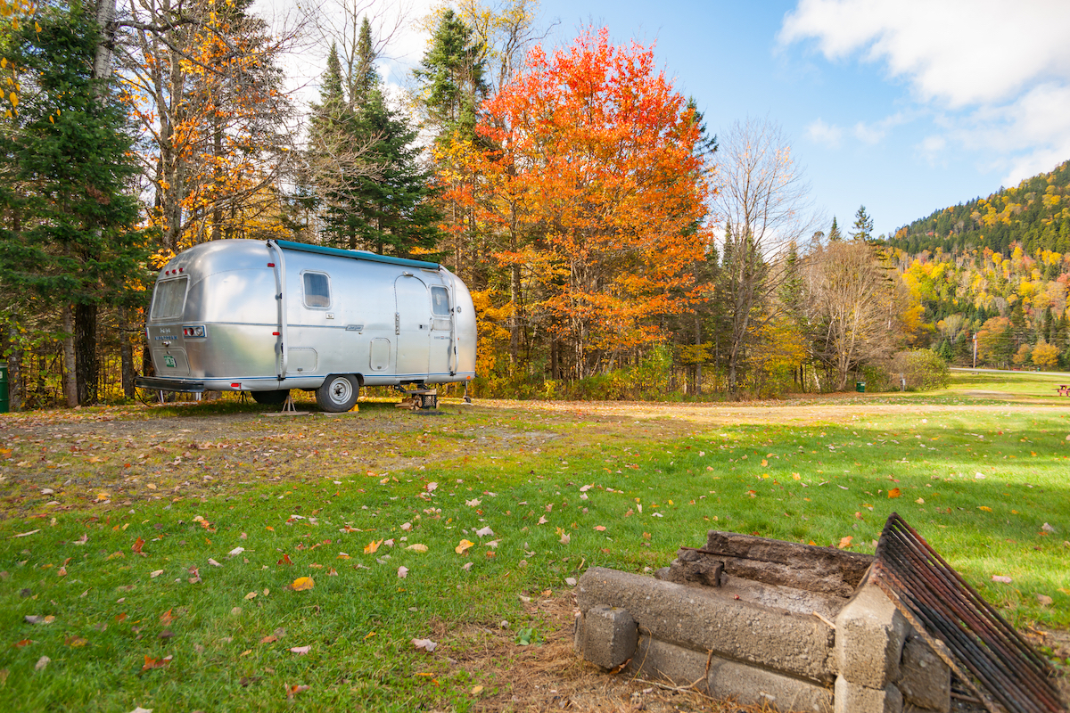 An Airstream travel trailer is located in front of trees turning orange.