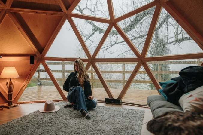 A young woman wearing autumn / winter clothes, inside a wooden dome with a geometric design in a forest.