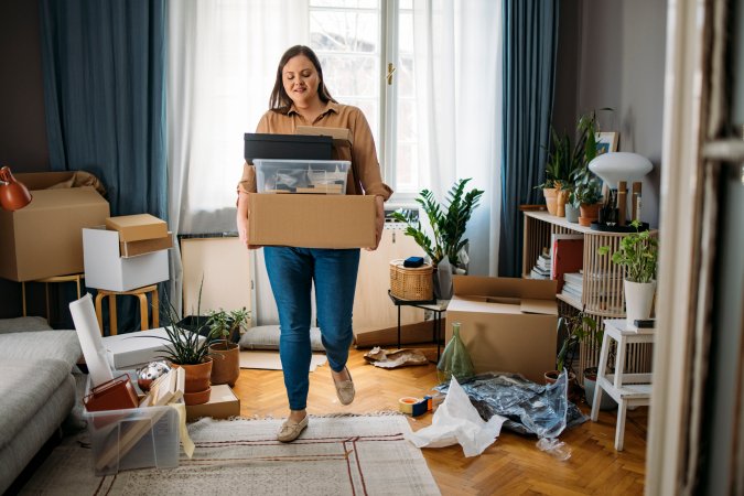 A woman is carrying boxes with personal belongings in a cluttered living room.