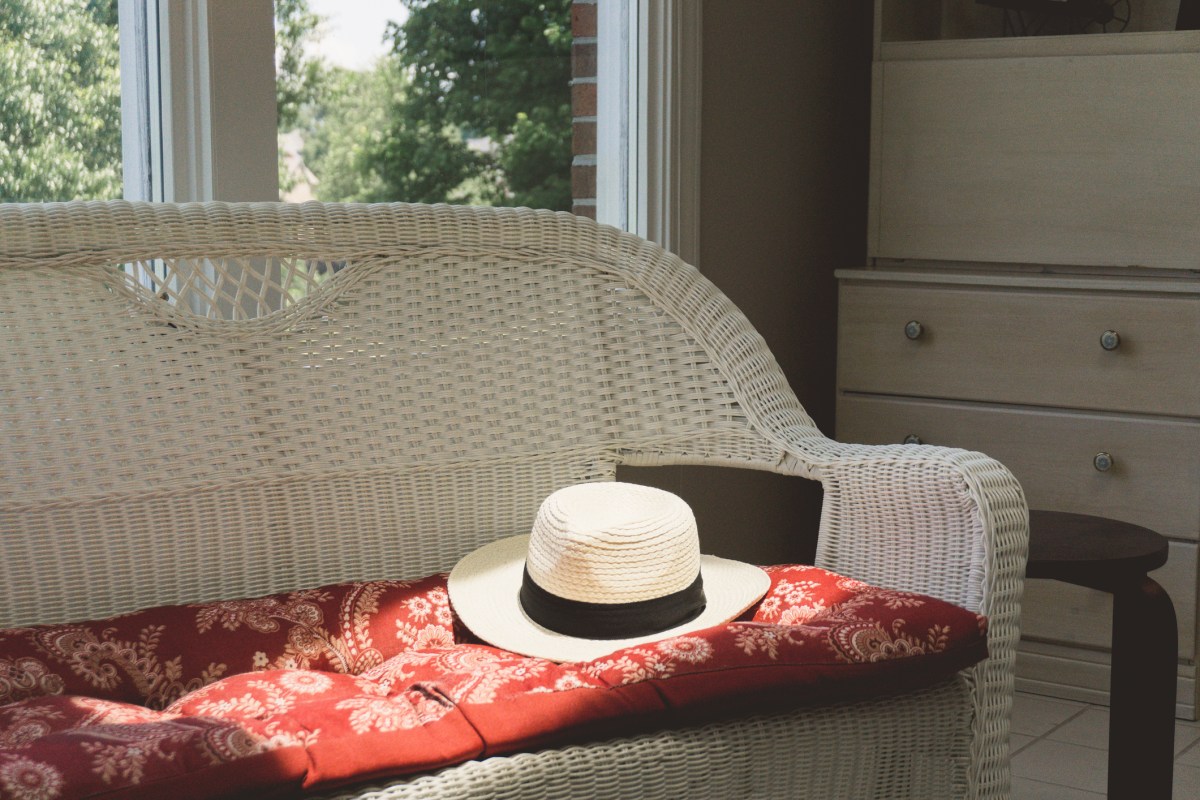 Summer day in the sunroom as a fedora hat rests on white wicker couch.